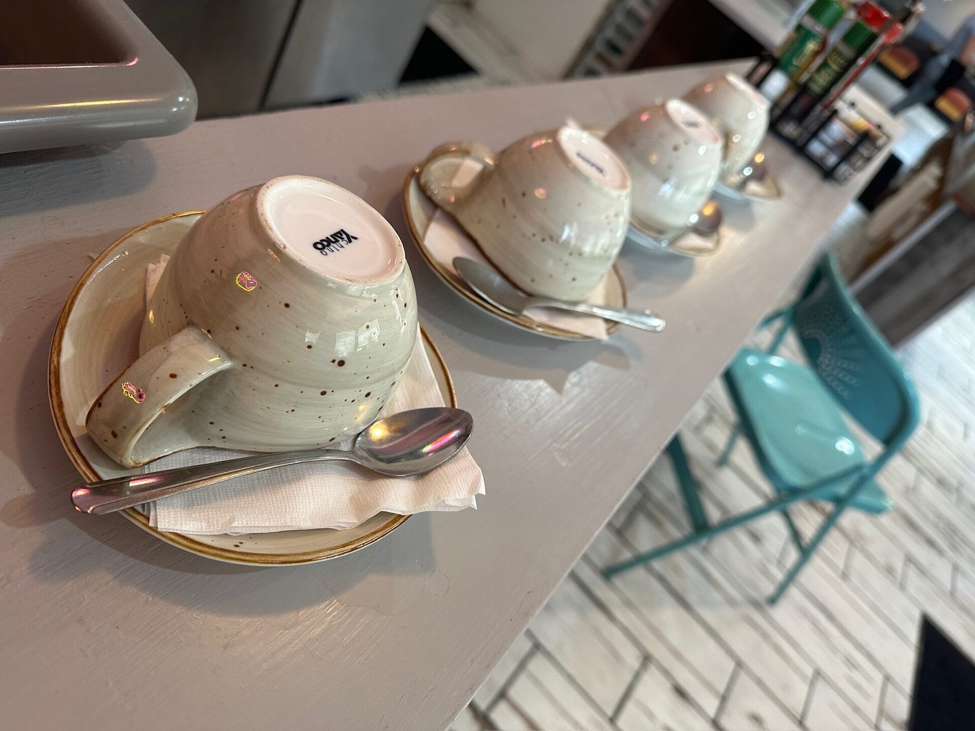 Coffee bar at Heavenly Diner & Donuts with speckled ceramic cups on the pale-pink counter and a teal chair